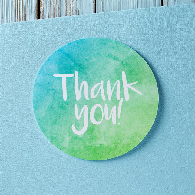 Autocollant vert brillant aquarelle Merci (Round thank-you sticker with a blue and green watercolor gradient and handwritten-style white text)