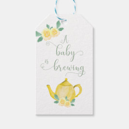 Ein Baby brät eine gelbe Teekanne mit Blumenbezug. Geschenkanhänger
