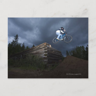 Ein Mountainbiker springt weg von einem Blockhaus Postkarte