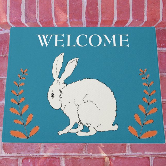 Elegantes Sitzen von Orangen-Pflanze aus weißem Ka Fußmatte (White sitting rabbit with tall orange plants on dark blue Welcome outdoor mat.)