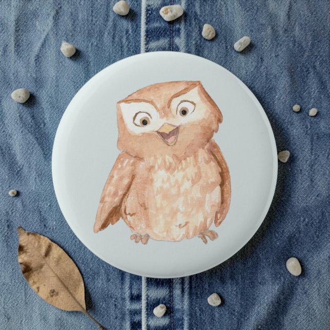 Happy Owl Wasserfarbe Button (A watercolor owl cartoon smiling on a pale blue pin, laid on denim amid pebbles and leaves.)