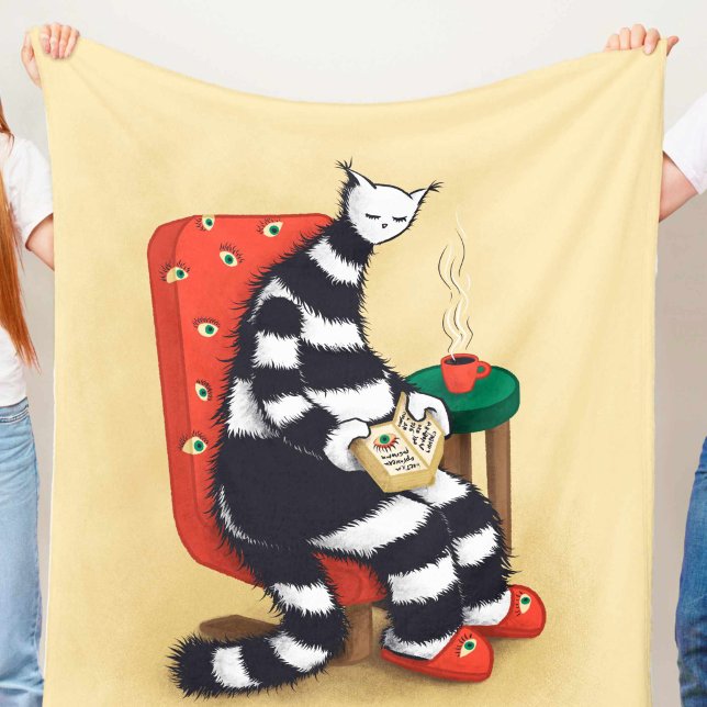 Lesen des Katalogbuches Fleecedecke (light beige blanket with weird reading cat character with book and coffee illustration held in hands)