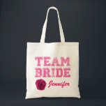 Pink Team Bride Tote Bag Tragetasche<br><div class="desc">Spaß Hochzeitsfeier Leinwand Tasche mit rosa Grafik Not Text lesen Team Bride. Benutzerdefinierter rosa Skripttext am unteren Rand ist bereit, für das gesamte Brautparty zu personalisieren. Die digitale Grafik einer rosa Rosenblüte schmückt den Boden links. Ideal, um alle wichtigen Dinge zu halten, die die Mitglieder des Brautparty nicht vergessen können....</div>