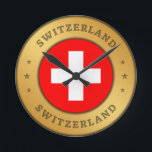 Schweizer Flagge Runde Wanduhr<br><div class="desc">Die Schweizer Fahne ist in einen gerundeten Goldring der Imitate eingeprägt. Das Wort "Schweiz" steht ganz oben und unten im Design und hat einen gepressten Effekt. Das Paar von drei dekorativen Sternen zueinander,  sind auch mit einem gepressten Effekt gestaltet.</div>