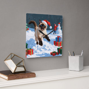 Siamkatze läuft im Schnee mit Weihnachtsmütze Quadratische Wanduhr