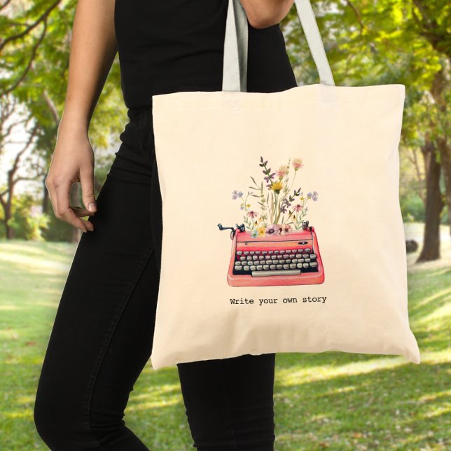 Tote Bag Écrivez votre propre histoire, Écrivains (A woman holding a tote bag with a typewriter graphic and wildflowers & text: "write your own story")